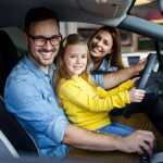 A father sits with his daughter behind the wheel of a car when a mother smiles after refinancing a car loan