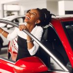 An African American woman smiles while holding on to the door of a bright red car.